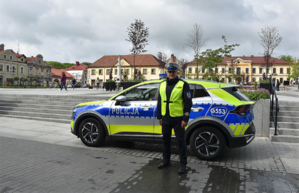 Policjant w kamizelce odbaskowej, stojący obok oznakowanego radiowozu na tle rynku w Bochni