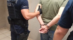 Zatrzymany z kajdankami zapiętymi z tyłu, a obok dwaj policjanci.