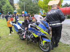 dziewczynka w policyjnej czapce siedzi na służbowym motocyklu obok stoi policjant a w tle uczestnicy pikniku