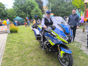 mężczyzna w czapce policjanta ruchu drogowego siedzi na policyjnym motocyklu, w tle uczestnicy pikniku