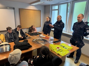 grupa uczniów na spotklaniu z policjantami. Na stołach znajdują się elementy wyposażenia policjantów oraz matreiały szkoleniowe
