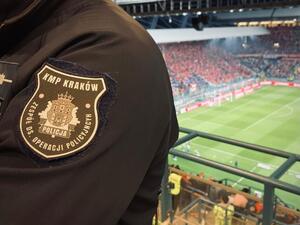 część policyjnego munduru, na którym znajduje się nszywka Zespołu do spraw Operacji Policyjnych. W oddali widać kibiców na trybunach