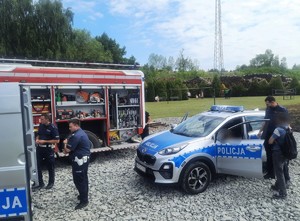Na zdjęciu wóz strażacki obok stoją dwaj policjanci. Po prawej radiowóz, stojący obok policjanta i mężczyzna.