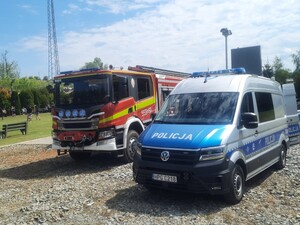 Na zdjęciu wóz strażacki i oznakowany radiowóz. W tle dzieci grające w piłkę.