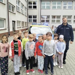 Umundurowany policjant stojący wspólnie z dziećmi przed oznakowanym radiowozem.