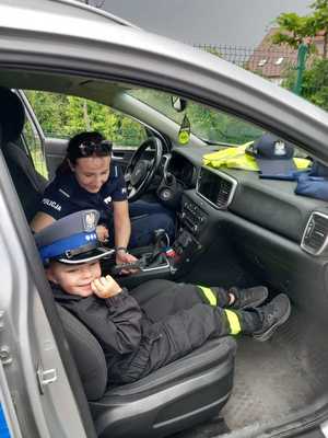 W radiowozie siedzi na miejscu kierowcy policjantka, a obok chłopiec, który ma założona policyjną czapkę.
