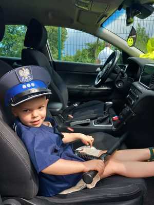 Mały chłopczyk ubrany w koszulę i czapke policyjną. Siedzi w radiowozie. W rekach trzyma tonfę.