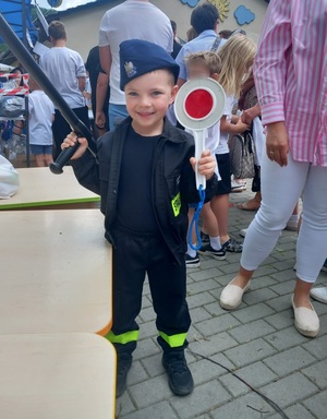 Mały chłopiec ubrany w strój strażaka. Na głowie ma policyjna czapkę i trzyma tarcze sygnalizacyjną. W tle osoby.