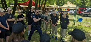 Grupa osób w czarnych koszulkach i czapeczkach z daszkiem oraz policjant prezentujący postawę strzelecką z bronią długą.