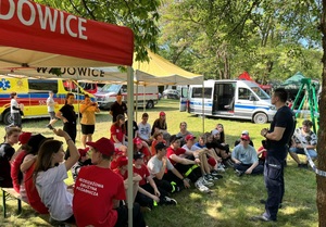 Umundurowany policjant, przed nimi grupa osób siedzących na ławkach. W tle samochody różnych służb mundurowych.