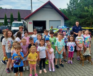 Grupa dzieci stojących przodem, za nimi policjant oraz w tle budynek i samochody.