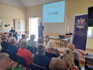 Sziedzący na sali seniorzy słuchają wypowiedzi policjanta.