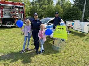 umundurowana policjantka stoi z dwójką dzieci przy stoisku profilaktycznym dziewczynki trzymają w ręce balon w kolorze granatowym