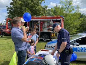 umundurowany policjant stoi przy stoisku profilaktycznym obok policjanta stoi mężczyzna trzymający dziecko na rękach
