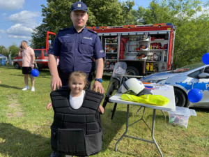 umundurowany policjant stoi z dziewczynką ubraną w kamizelkę policyjną na tle samochodu straży pożarnej