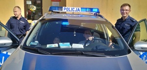 Na zdjęciu po prawej i lewej stronie stoi umundurowany policjant. W radiowozie siedzie dwóch chłopców, z których jeden ma założona policyjna czapkę.