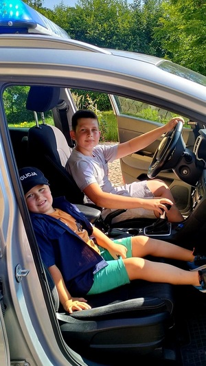 W policyjnym radiowozie, w którym są pootwierane drzwi z przodu, siedzą dwaj chłopcy. Ten, który siedzi na miejscu pasażera ma założoną czapkę i koszule policyjną.