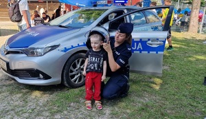 policnatka zakładająca policyjny kask na głowę małego chłopca stojącego przy radiwozie. Wokół radiwozu widać też inne osoby uczestniczące w pikniku