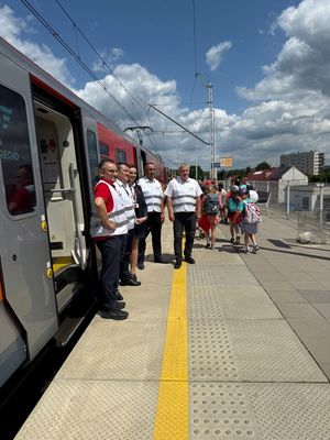 osoby obsługujące przejazd w kamizelkach odblaskowych stoją przy pociągu na peronie. Obok widać przechodzące dzieci