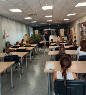 Na zdjęciu policjantka, przed nią grupa dzieci siedzących w ławkach, są tyłem. Osoby są w pomieszczeniu biblioteki.