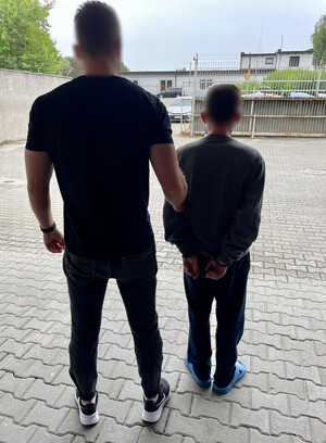 nieumundurowany policjant prowadzi na policyjnym parkingu zatrzymanego mężczyznę z założonymi kajdankami na ręce trzymane z tyłu przed