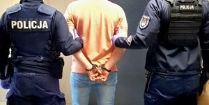Zatrzymany mężczyzna, który ma założone kajdanki na rękach trzymanych z tyłu. Po prawej i lewej stronie są umundurowani policjanci. Osoby są tyłem.