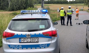 Po lewej radiowóz, po prawej dwóch policjantów ruchu drogowego i mężczyzna w pomarańczowej kamizelce odblaskowej stojących tyłem.  Na drodze dron. W tle samochód rajdowy.