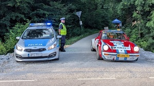 Po lewej stronie radiowóz obok stoi policjant ruchu drogowego. Po prawek samochód rajdowy.