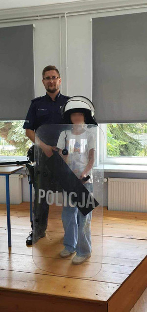 Umundurowany policjant oraz dziecko mające na sobie policyjny kask oraz trzymające tarczę z napisem POLICJA