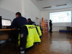 Policjant prowadzący szkolenie, stojąc przy ścianie gdzie wyświetlany jest plan szkicu miejsca zdarzenia drogowego, tłumaczy sposób wykonywania zadań. Na Sali znajdują się policjanci siedzący przy swoich stanowiskach komputerowych.