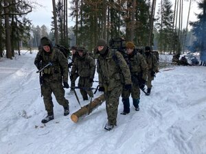 Grupa uczestników niesie wspólnie kłodę drewna podpierając ją na paskach karabinów