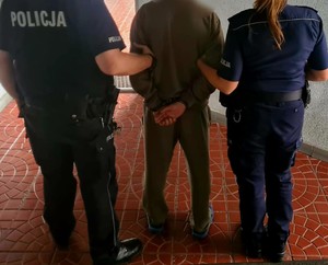 Po lewej stronie umundurowany policjant, po prawej policjanta, a po środku zatrzymany mężczyzna, który ma założone kajdanki na ręce trzymane z tyłu.