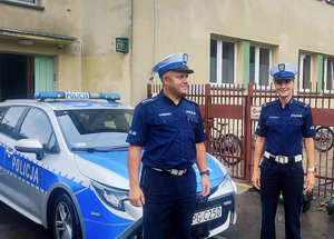 policjant i policjantka przed budynkiem szkoły, za nimi zaparkowany radiowóz