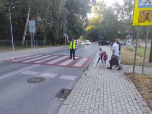 Policjant stoi na przejściu dla pieszych i daje kierującym sygnał lizakiem aby sie zatrzymali bo przez przejście przechodzą dzieci.