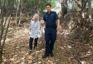 Umundurowany policjant prowadzący pod rekę starsza kobietę. Osoby są w lesie.