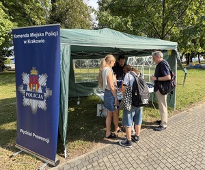 Policjant na stanowisku profilaktycznym. Przy stoisku rodzina patrzy na materiały.