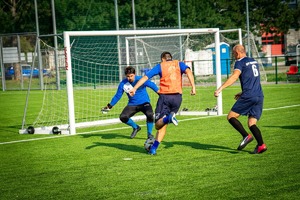 Przed bramką dwóch piłkarzy z których jeden kopie w piłkę, przed nim bramkarz