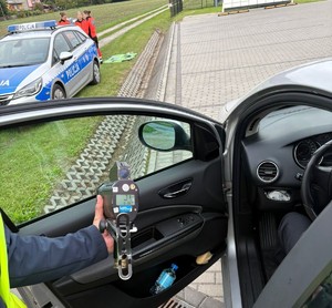 Kampania Mistrzu zwolnij i Road Safety Days  policjant z miernikiem prędkości przy samochodzie w tle radiowóz i ratownicy medyczni