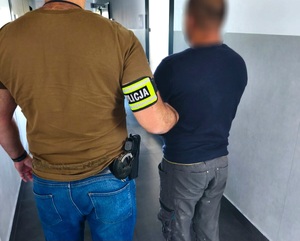 Na zdjęciu po lewej stronie policjant w stroju cywilnym z bronią i opaską z napisem Policja na prawej ręce, który trzyma zatrzymanego mężczyznę.