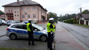 Policjanci stoją koło radiowozu i kontrolują prędkość jadących drogą pojazdów
