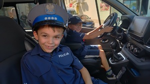 Dwóch chłopców siedzących w radiowozie w koszulach i czapkach policyjnych.