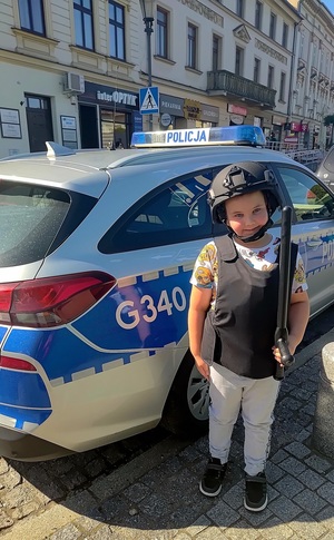 Na tle radiowozu chłopiec ubrany w elementy wyposażenia policyjnego.