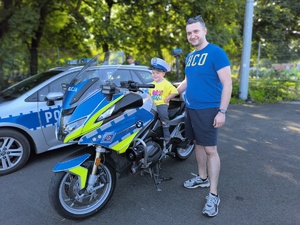 dziecko na policyjnym motocyklu obok stoi opiekun dziecka, w tle radiowóz