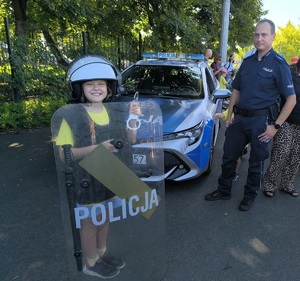 dziecko ubrane w elementy policyjnego umundurowania pozuje do zdjęcia z policjantem przy radiowozie