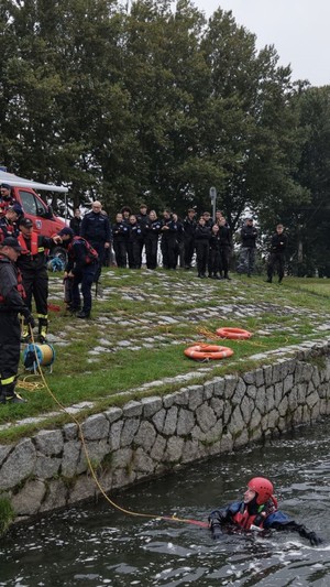 Uczniowie i policjanci stojący wraz zze strazakami nad brzegike w wodzie widoczny strazak