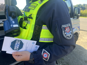 policjant trzyma w ręce radarowy miernik prędkości i ulotkę zwolnij mistrzu