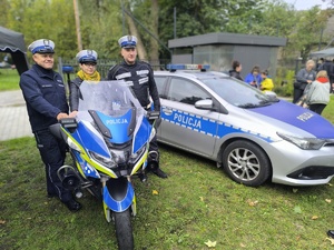 Umundurowani policjanci w towarzystwie kobiety siedzącej na policyjnym motocyklu. Obok widać oznakowany radiowóz