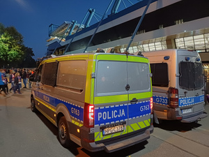 Dwa radiwozy zaparkowane przy stadionie obok, których przechodzą kibice udający się na mecz