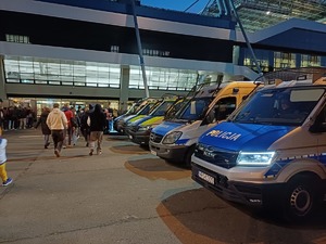 radiowozy zaparkowane przy stadionie, przed którymi przechodzą osoby udające się na mecz