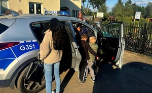 Na zdjęciu radiowóz i osoby w tym dzieci go oglądające. W tle budynek.